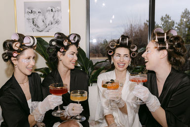 Bride and bridesmaids in robes with hair rollers and under-eye patches, laughing and toasting colorful cocktails during a wedding morning getting-ready session