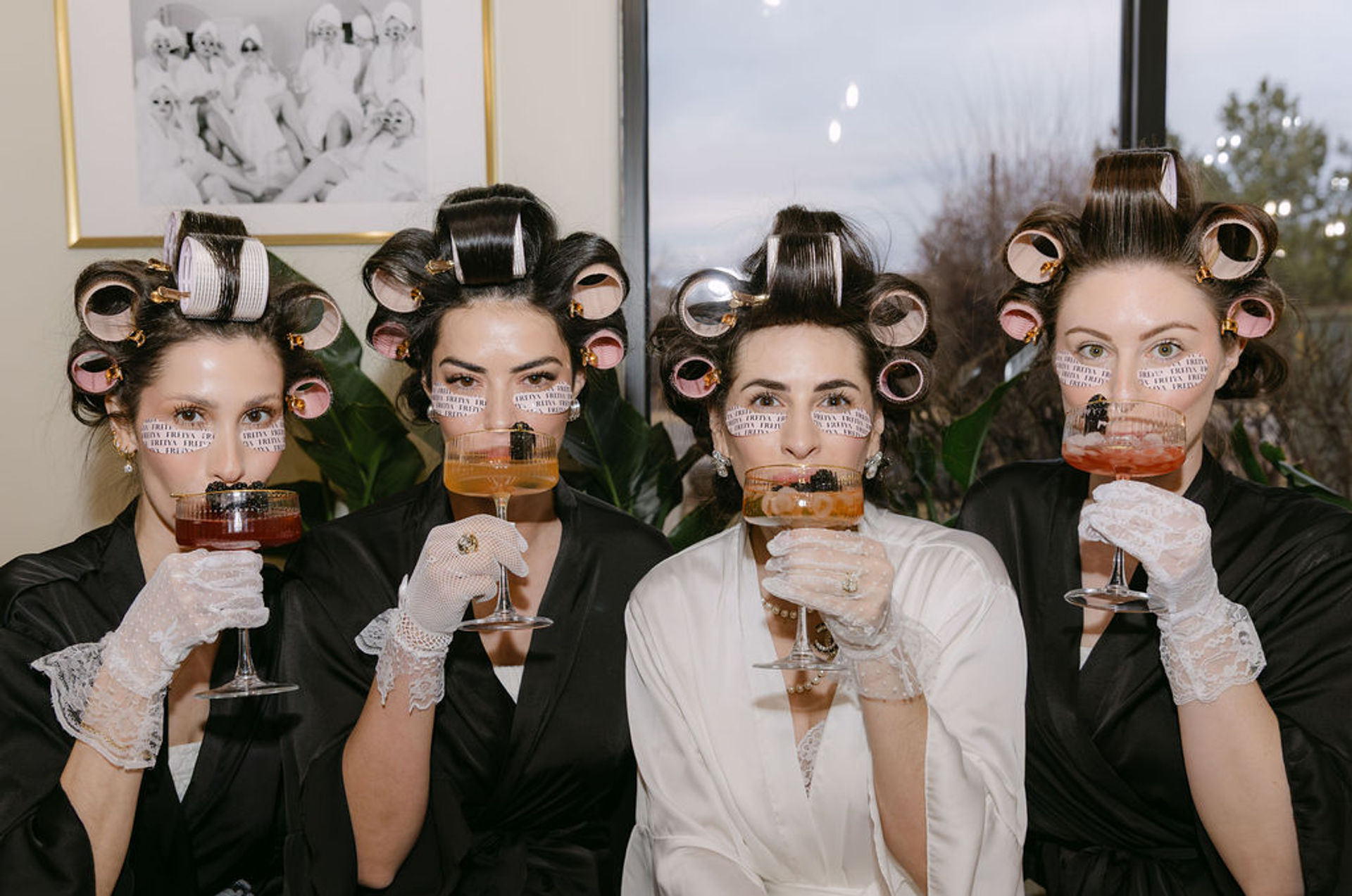 Four women in robes with hair rollers, under-eye patches and lace gloves sipping cocktails indoors in a playful bridal-party getting-ready scene