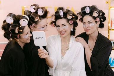 Four women in a beauty salon wearing robes, large hair rollers and under-eye patches, smiling playfully into a handheld mirror during a spa-style styling session.