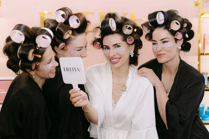 Four women in a beauty salon wearing robes, large hair rollers and under-eye patches, smiling playfully into a handheld mirror during a spa-style styling session.