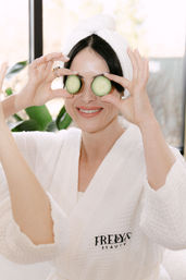 Woman in a white spa robe and towel wrap smiling while holding cucumber slices over her eyes in a bright at-home spa setting
