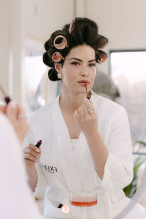 Person in a white robe applying pink lip gloss at a vanity mirror, hair in pink rollers, holding the applicator and bottle, with a cocktail glass on the counter — cozy at-home beauty routine.