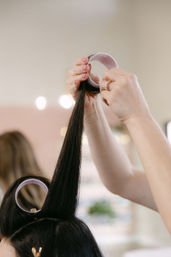Close-up of a stylist’s hands rolling long dark hair into foam curlers in a bright salon, showing sectioned strands and styling process.