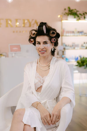 Smiling woman in a white robe and lace bralette with hair in rollers and under-eye patches, seated in a pastel beauty salon interior for hair and makeup prep.