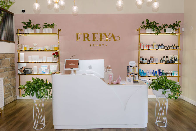 Bright boutique beauty reception with a white curved front desk, pink accent wall, gold shelving stocked with skincare products, globe pendant lights, and green potted plants.