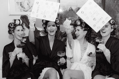 Black-and-white image of four women in robes and hair rollers laughing and toasting with champagne in a bridal suite, wearing under-eye patches and lace gloves while tossing wedding cards.