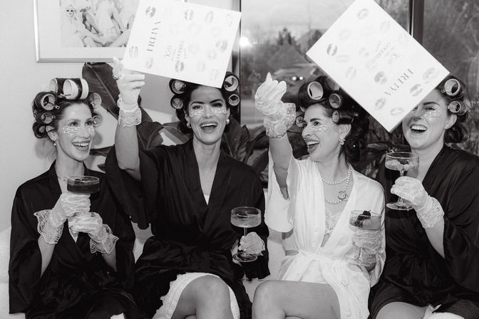Black-and-white image of four women in robes and hair rollers laughing and toasting with champagne in a bridal suite, wearing under-eye patches and lace gloves while tossing wedding cards.