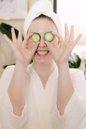 Smiling person in a white bathrobe and towel turban holding cucumber slices over their eyes for a playful home spa skincare moment