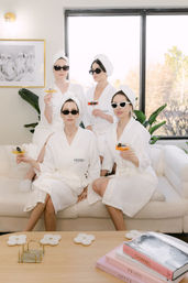 Four women in white spa robes and towel turbans wearing sunglasses, sipping cocktails while lounging on a sunlit modern living-room sofa with plants and a coffee table stacked with books.