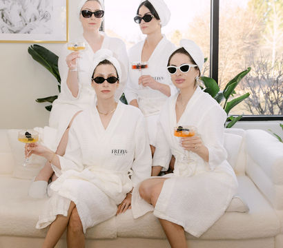 Four women in white spa robes and towel turbans wearing sunglasses, sipping cocktails while lounging on a sunlit modern living-room sofa with plants and a coffee table stacked with books.