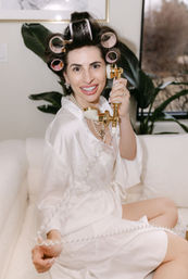 Smiling woman in a white satin robe and pearl necklace with pink hair rollers, playfully holding a gold vintage telephone handset while seated on a cream sofa in a cozy indoor setting.