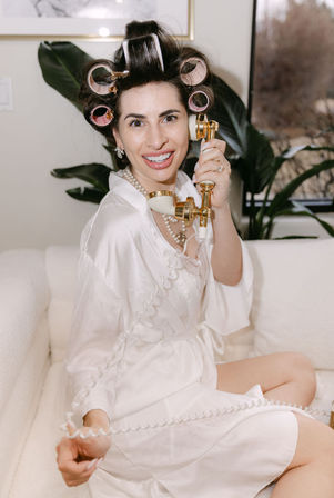 Smiling woman in a white satin robe and pearl necklace with pink hair rollers, playfully holding a gold vintage telephone handset while seated on a cream sofa in a cozy indoor setting.