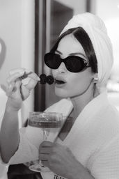 Black-and-white close-up of a glamorous woman in a towel turban and robe, wearing dark sunglasses, nibbling blackberries on a skewer while holding a champagne coupe — spa-style luxury vibe.