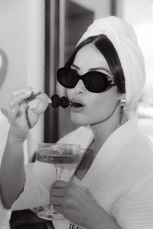 Black-and-white close-up of a glamorous woman in a towel turban and robe, wearing dark sunglasses, nibbling blackberries on a skewer while holding a champagne coupe — spa-style luxury vibe.