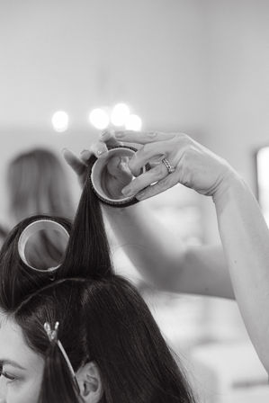 Black-and-white close-up of a hairstylist rolling dark hair into a velcro roller in a salon, hand with a ring and hair clipped and sectioned for styling.