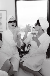 Black-and-white photo of two friends in white spa robes and towel turbans wearing sunglasses, laughing and eating cherries while holding champagne coupes during a playful indoor spa day.