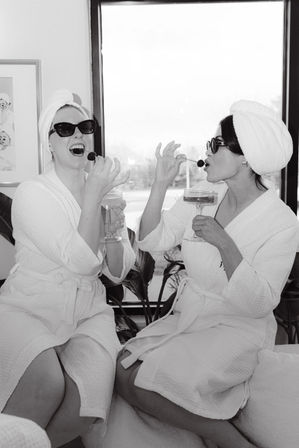 Black-and-white photo of two friends in white spa robes and towel turbans wearing sunglasses, laughing and eating cherries while holding champagne coupes during a playful indoor spa day.