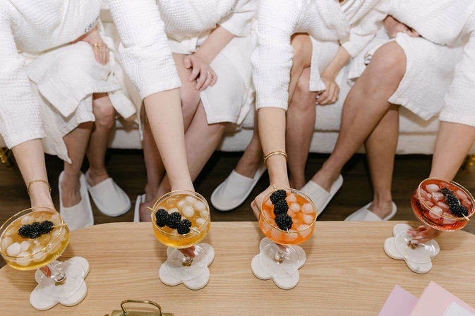 Four people in white spa robes and slippers sit on a couch, each reaching for a colorful coupe glass of iced cocktail topped with blackberries on a wooden table with flower-shaped coasters.