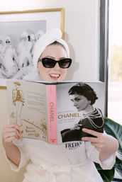 Smiling person in a white bathrobe with towel turban and sunglasses, relaxing by a sunny window and reading a Chanel fashion biography for a chic at-home spa day.