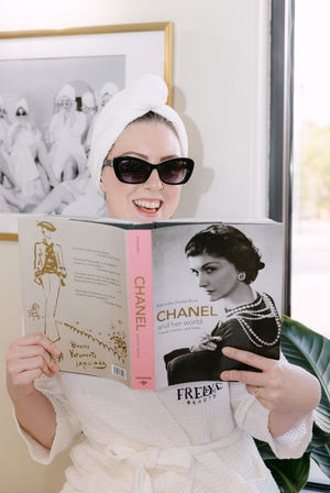 Smiling person in a white bathrobe with towel turban and sunglasses, relaxing by a sunny window and reading a Chanel fashion biography for a chic at-home spa day.