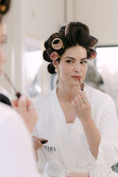 Woman in a white waffle robe with pink hair rollers applying rosy lip gloss at a vanity mirror — relaxed at-home getting-ready beauty routine.