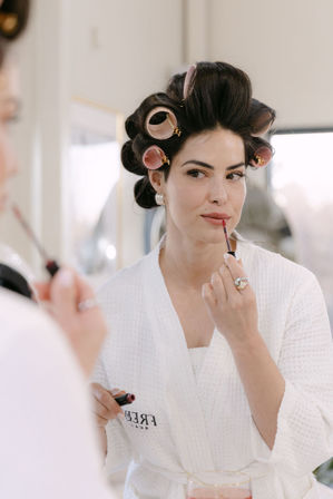 Woman in a white waffle robe with pink hair rollers applying rosy lip gloss at a vanity mirror — relaxed at-home getting-ready beauty routine.