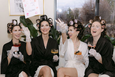 Four women in hair rollers and satin robes, wearing under-eye patches and lace gloves, laughing on a couch and toasting with cocktails in a bright getting-ready lounge.