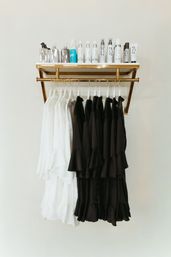 Gold wall-mounted shelf topped with haircare bottles above a neat row of white and black ruffled dresses on hangers — minimalist boutique clothing display