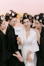 Bridal party getting ready in a salon-style suite: four women in hair rollers and under-eye patches wearing robes, laughing as the bride in white holds a handheld mirror for a selfie.