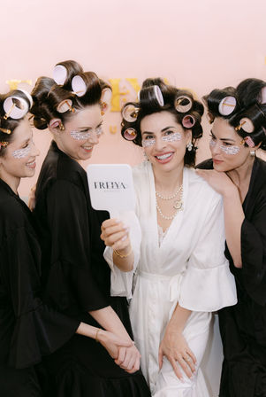 Bridal party getting ready in a salon-style suite: four women in hair rollers and under-eye patches wearing robes, laughing as the bride in white holds a handheld mirror for a selfie.