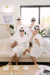 Four women in white spa robes and towel turbans wearing sunglasses, seated on a cream sofa in a bright lounge, raising cocktail glasses and smiling near a large window and plants.