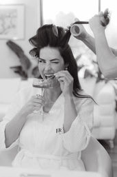 Black-and-white image of a woman in a spa robe playfully winking and sipping champagne while a stylist rolls her hair in a salon