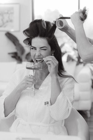 Black-and-white image of a woman in a spa robe playfully winking and sipping champagne while a stylist rolls her hair in a salon
