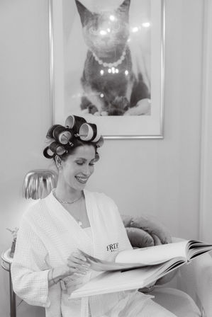Smiling woman in a waffle robe with hair rollers reading a large book in a cozy salon setting, framed cat portrait visible on the wall behind her.