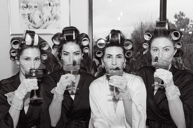 Black-and-white photo of four women in hair rollers, silk robes and lace gloves sipping cocktails while wearing under-eye patches during a bridal-party getting-ready moment
