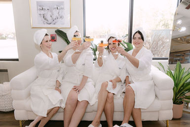 Four friends in white spa robes and towel turbans toasting colorful cocktails on a plush couch during a bright spa-day celebration with plants and large windows.