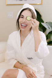 Smiling woman in a white waffle robe and towel turban holding a cucumber slice over one eye in a bright at-home spa setting with a leafy plant in the background.