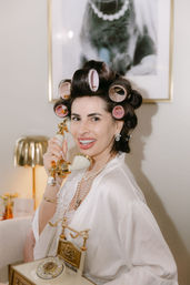 Smiling woman in a satin robe with pink hair rollers and pearl necklace holding a cream-and-gold vintage rotary phone at a chic home vanity.