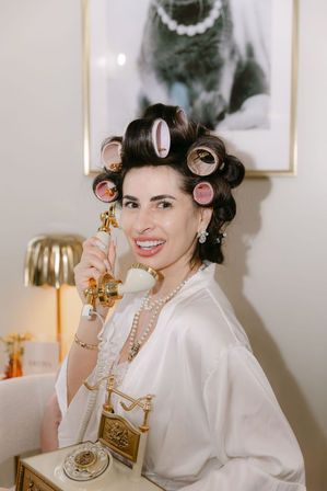 Smiling woman in a satin robe with pink hair rollers and pearl necklace holding a cream-and-gold vintage rotary phone at a chic home vanity.