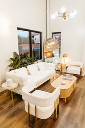 Cozy chic modern living room with cream boucle sofa and matching armchairs with gold legs, wooden oval coffee table, tall arched floor mirror, indoor plants by black-framed windows, brass chandelier and warm hardwood floors.