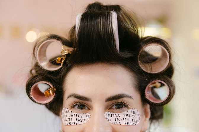 Close-up of a woman in a beauty salon with dark hair in pink and beige hair rollers and under-eye patches, prepping for styling.
