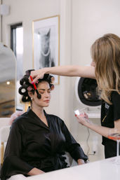 Makeup artist applies blush with a brush to a seated woman in hair rollers wearing a black satin robe at a beauty salon.