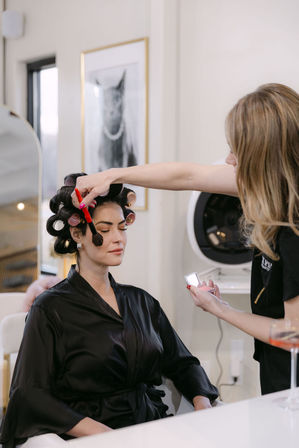 Makeup artist applies blush with a brush to a seated woman in hair rollers wearing a black satin robe at a beauty salon.