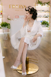 Woman in a white robe with hair rollers and under-eye patches sitting in a modern beauty salon chair, wearing pearl jewelry and white open-toe heels amid plants and product shelves.