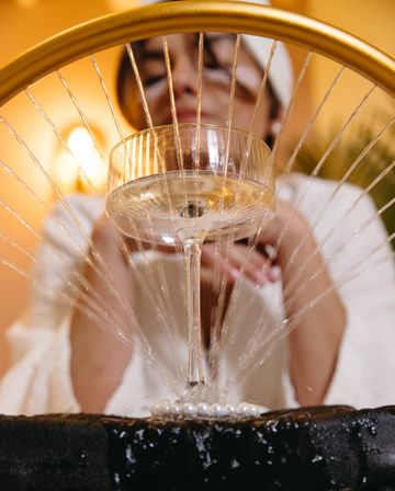 Close-up of a champagne coupe in a decorative fountain — sparkling water jets radiate beneath a gold hoop with warm indoor lighting and a blurred person in a white robe in the background.