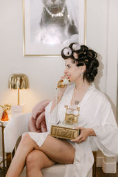 Person in white robe with hair rollers and pearl necklace, smiling while holding an ornate gold vintage telephone in a cozy vanity chair beside a lamp and framed cat print.