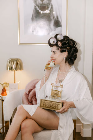 Person in white robe with hair rollers and pearl necklace, smiling while holding an ornate gold vintage telephone in a cozy vanity chair beside a lamp and framed cat print.