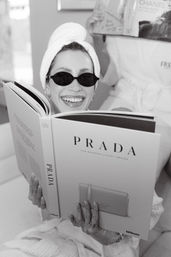 Black-and-white photo of a smiling woman in a towel turban and small sunglasses reading a large luxury fashion coffee-table book while wearing a robe and rings in a relaxed indoor spa-style setting, with another person reading in the background.