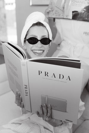 Black-and-white photo of a smiling woman in a towel turban and small sunglasses reading a large luxury fashion coffee-table book while wearing a robe and rings in a relaxed indoor spa-style setting, with another person reading in the background.