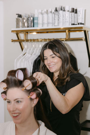 Hairstylist smiling as she sets pink hot rollers in a client's hair in a bright beauty salon with a shelf of hair products
